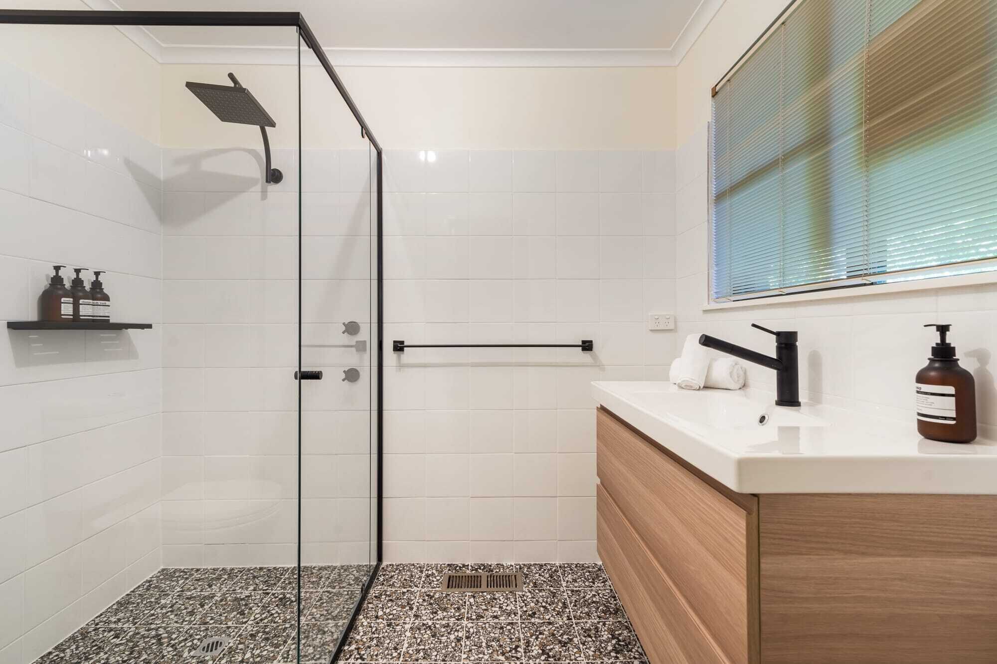 A modern bathroom with a glass shower enclosure, a white tiled wall, a wooden vanity, and a window with blinds.