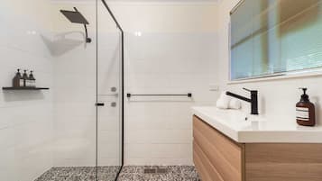 A modern bathroom with a glass shower enclosure, a white tiled wall, a wooden vanity, and a window with blinds.