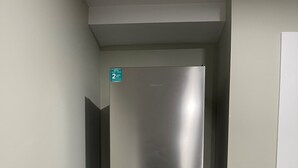 Un réfrigérateur en acier inoxydable se trouve dans un couloir étroit, avec une porte blanche et un petit panneau vert en haut.