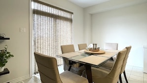 Un coin repas comprenant une table à plateau en verre, six chaises de couleur beige, un lustre et une fenêtre avec des stores.