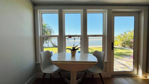 Ein Zimmer mit einem runden Tisch und zwei Stühlen, große Fenster mit Blick auf den Strand und Palmen.