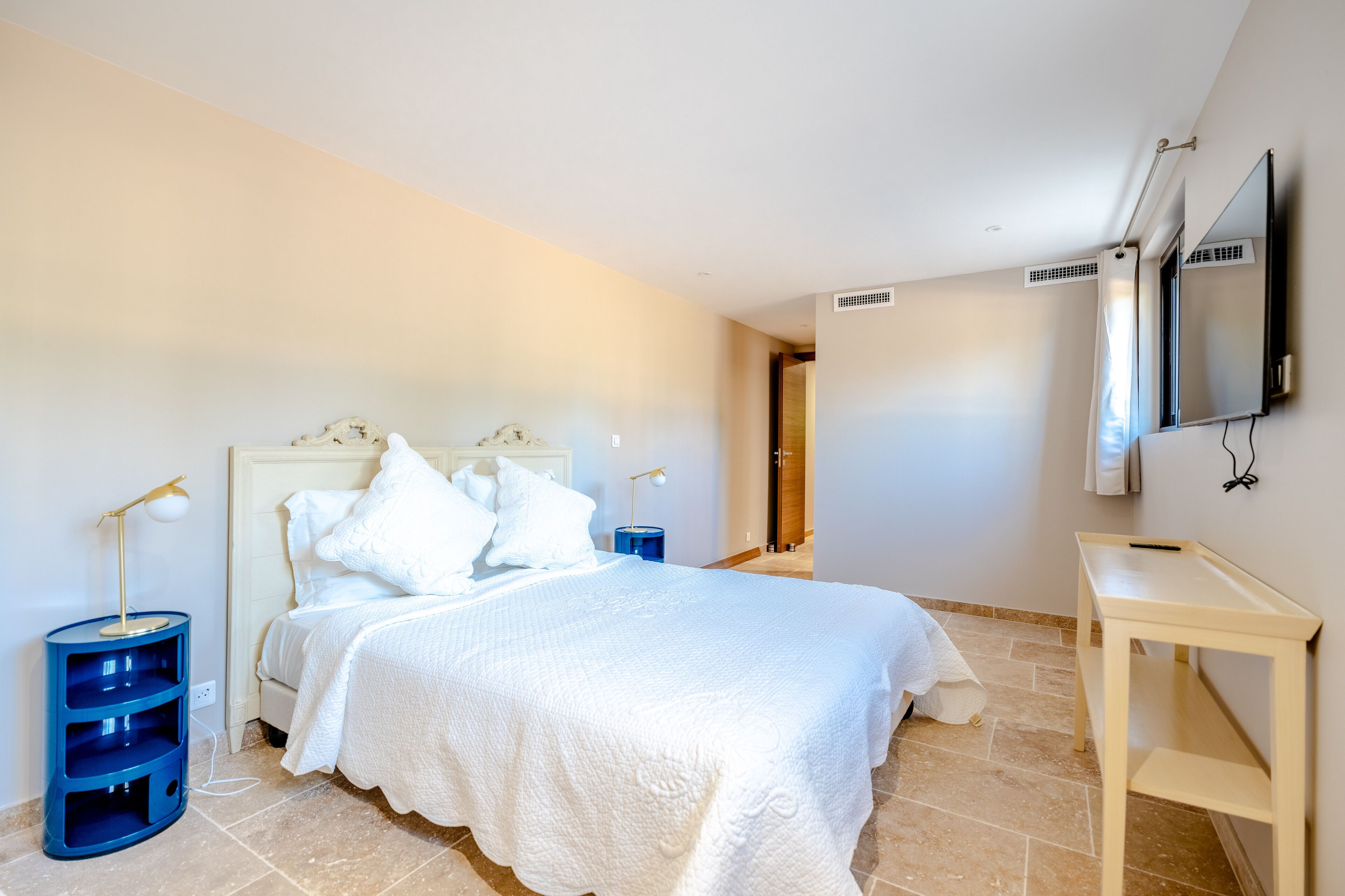 A bedroom with a large bed, a bedside table, a TV mounted on the wall, and a door leading to another room.