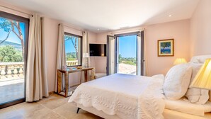 A bedroom with a large bed, a TV, a desk, and a balcony with a view of the landscape. - Magnificient Villa totally renovated in 2023 with sea view and Saint Tropez   (Grimaud)