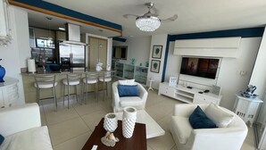 A modern living room with a TV, white sofas, a coffee table, and a dining area with chairs. - Amazing 2BR Beach Apartment with Ocean View for 6 (Playa Juan Dolio)