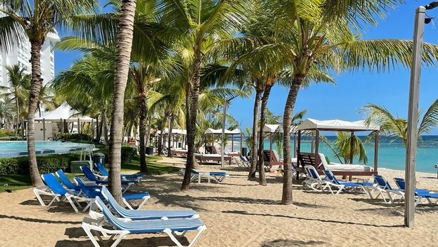 A beachside resort with sun loungers, palm trees, and a clear view of the ocean. - Amazing 2BR Beach Apartment with Ocean View for 6 (Playa Juan Dolio)