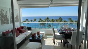 A modern living room with a balcony overlooking the ocean, furnished with a sofa, dining table, and chairs. - Amazing 2BR Beach Apartment with Ocean View for 6 (Playa Juan Dolio)