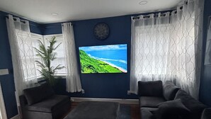 A modern living room with a large flat-screen TV mounted on the wall, two sofas, a coffee table, and a potted plant. - The Blue backyard studio suite!!  Convenience and practicality in one place!! (Allentown)