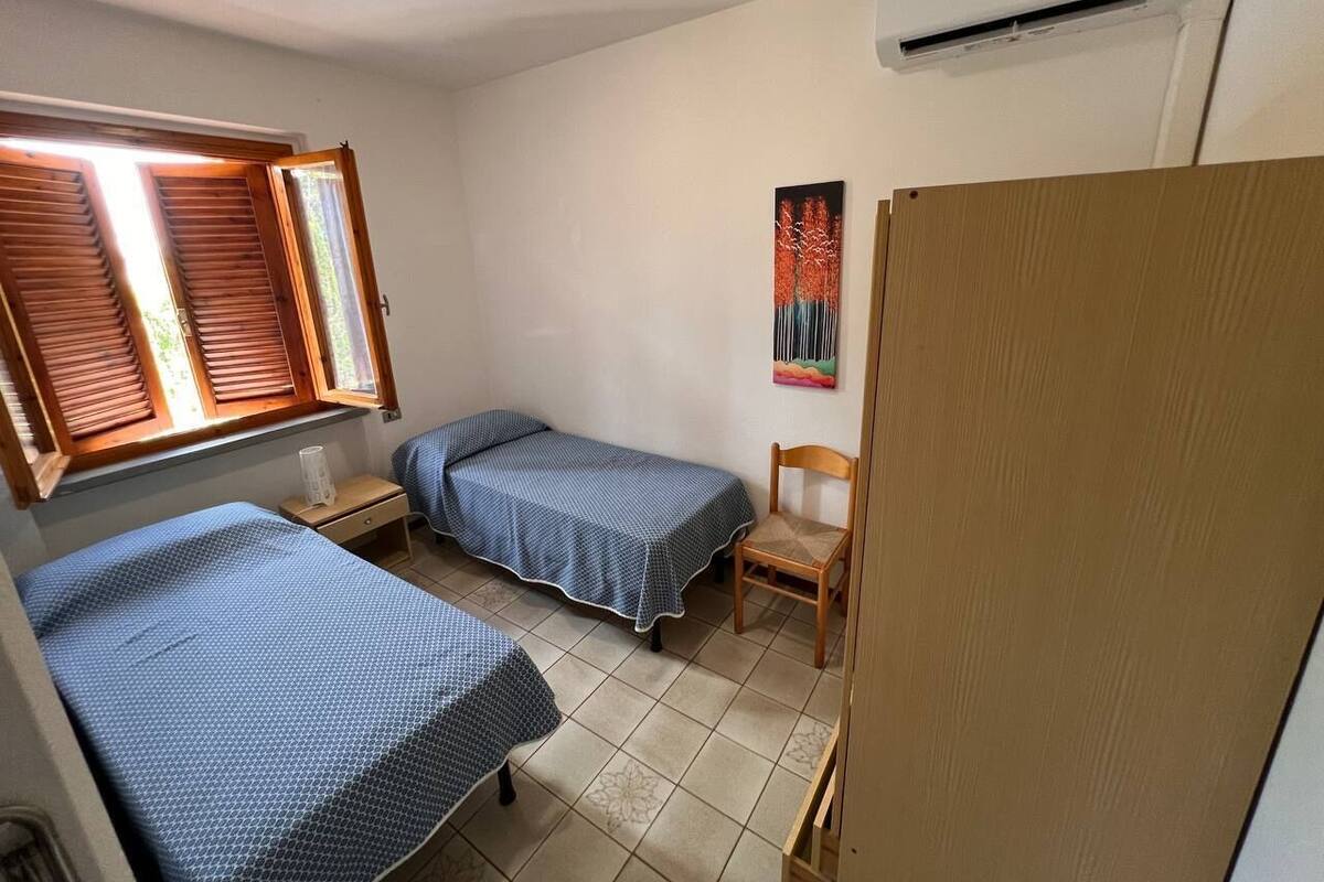 A bedroom with two beds, a chair, a nightstand, a window with blinds, and a wall-mounted air conditioner.