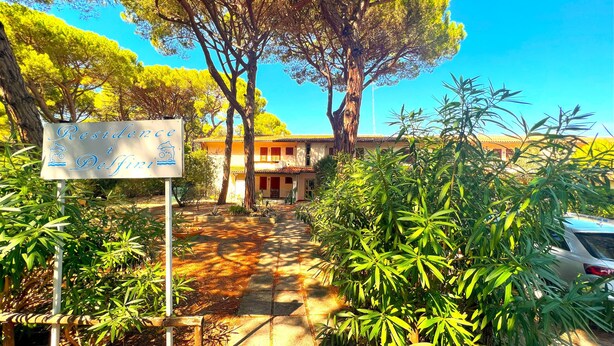 A sign for 'Residence Delfini' with a pathway leading to a building surrounded by trees and plants.