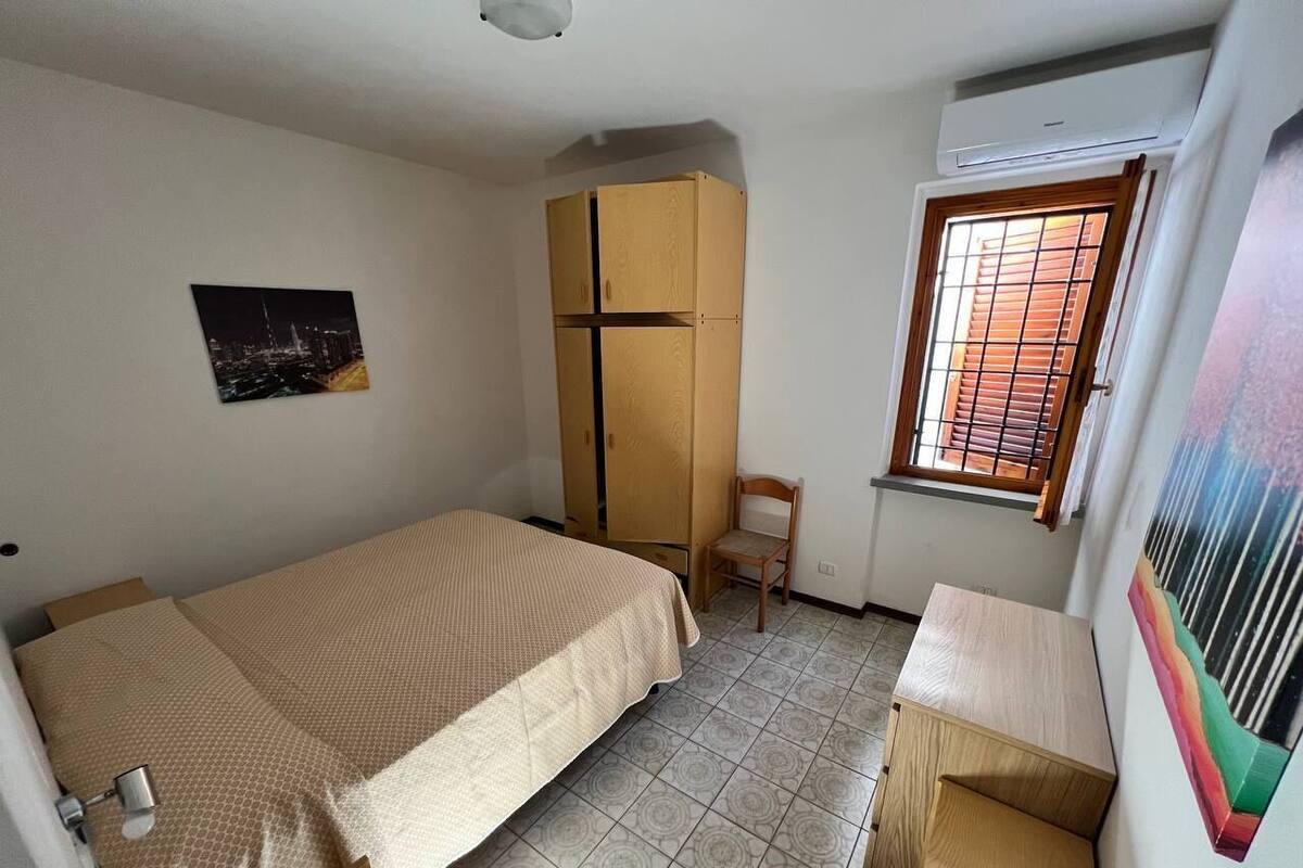 A small, single-room apartment with a bed, a desk, a chair, a wardrobe, and a window.