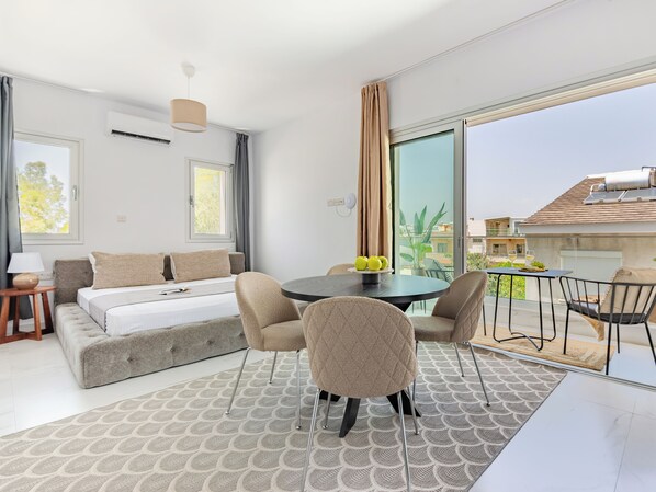 A modern hotel room with a sofa bed, a dining table with chairs, a large window, and a balcony with a view of the city.