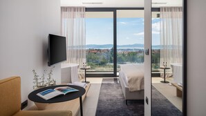 A modern hotel room with a large sliding glass door leading to a balcony with a city view.
