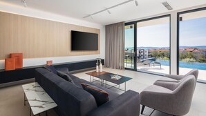 A modern living room with a large flat-screen TV, a sofa, a coffee table, and a view of a swimming pool and buildings.