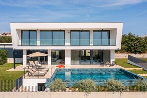 Une maison moderne à deux étages, dotée d’une piscine, de grandes fenêtres et d’un espace salon extérieur.