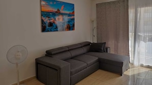 Una sala moderna con un sofá seccional gris, un ventilador de pie, un cuadro de una escena costera y un ventanal con cortinas traslúcidas.