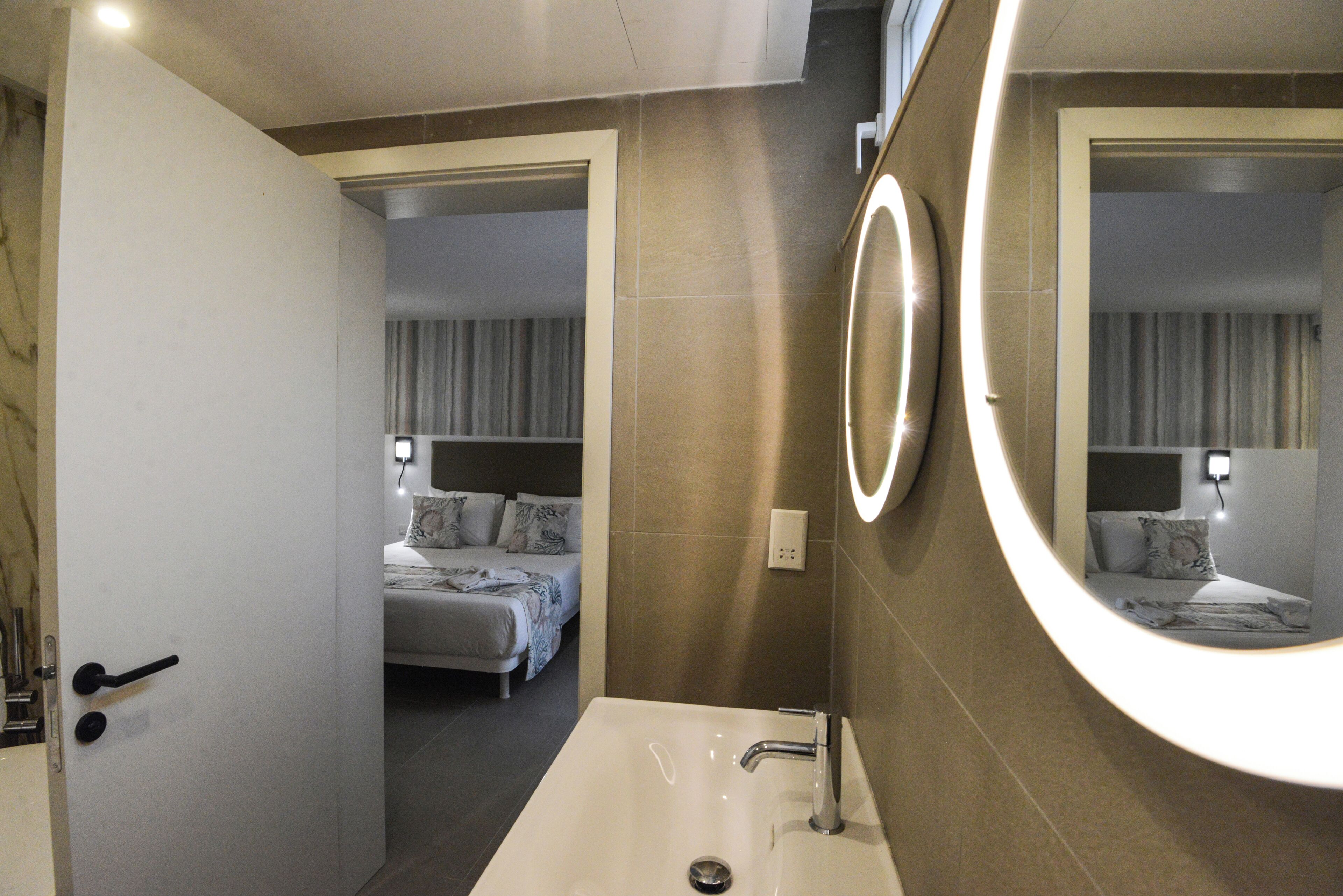 A modern bathroom with a large mirror, a sink, and a door leading to a bedroom.