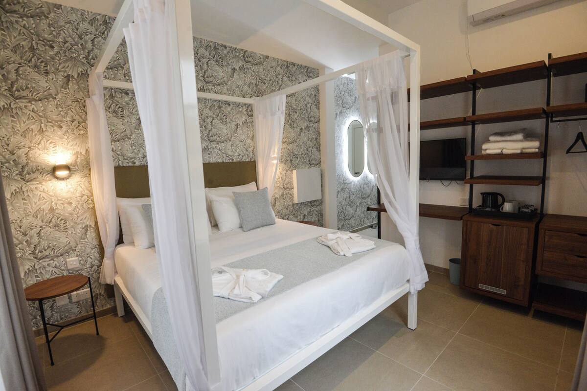 A four-poster bed with white bedding, a bedside table, a wall-mounted TV, and a wooden cabinet with shelves.