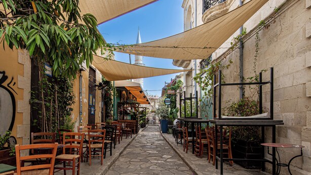 En smal kullerstensgata med uteservering, trästolar och bord under skuggiga områden.