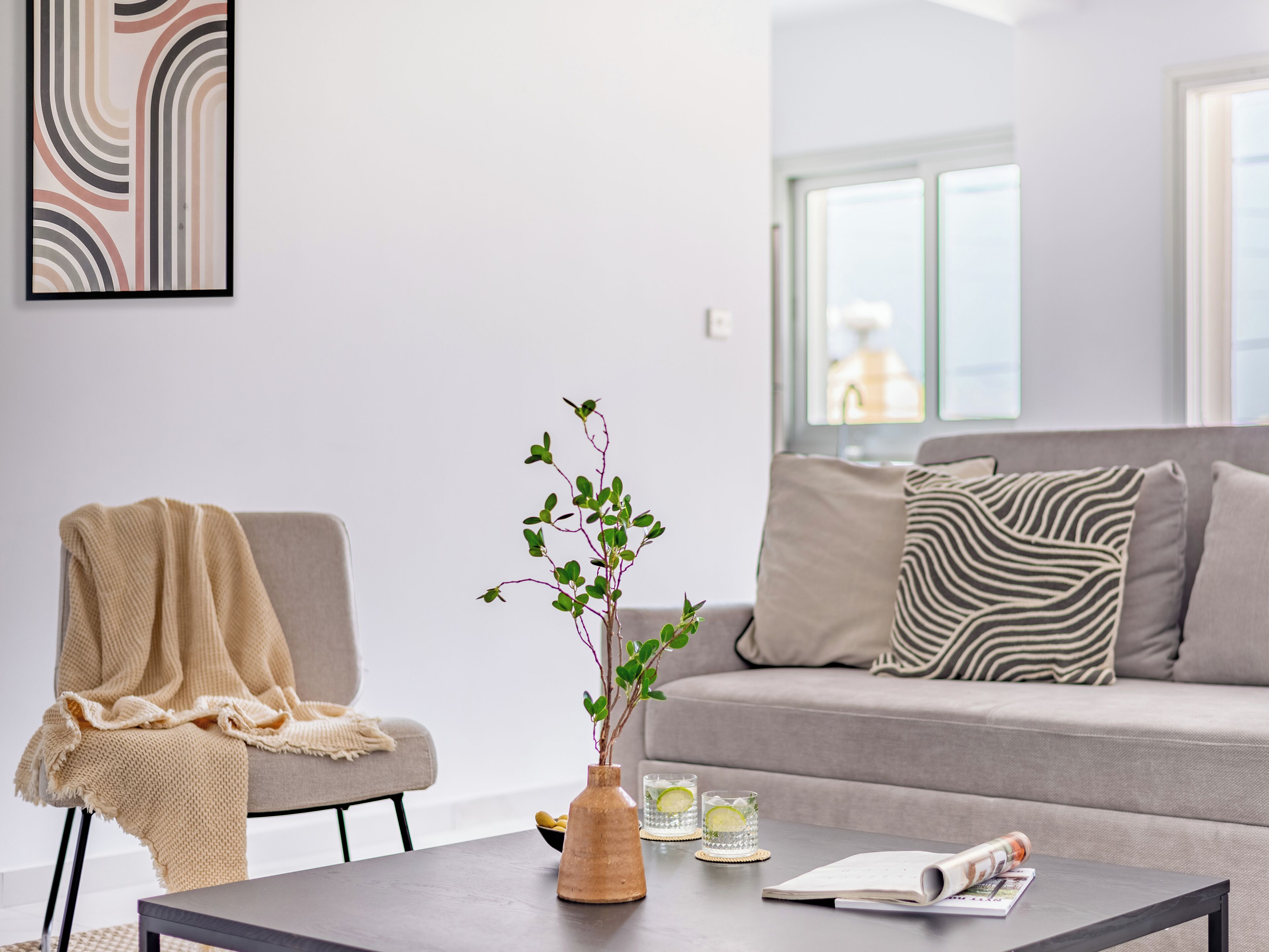 Una sala de estar moderna con un sofá, una mesa de centro, una silla con una manta, un jarrón con una planta y un cuadro en la pared.