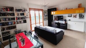 A modern living room with a glass dining table, a black sofa, and a bookshelf filled with books and decorative items.