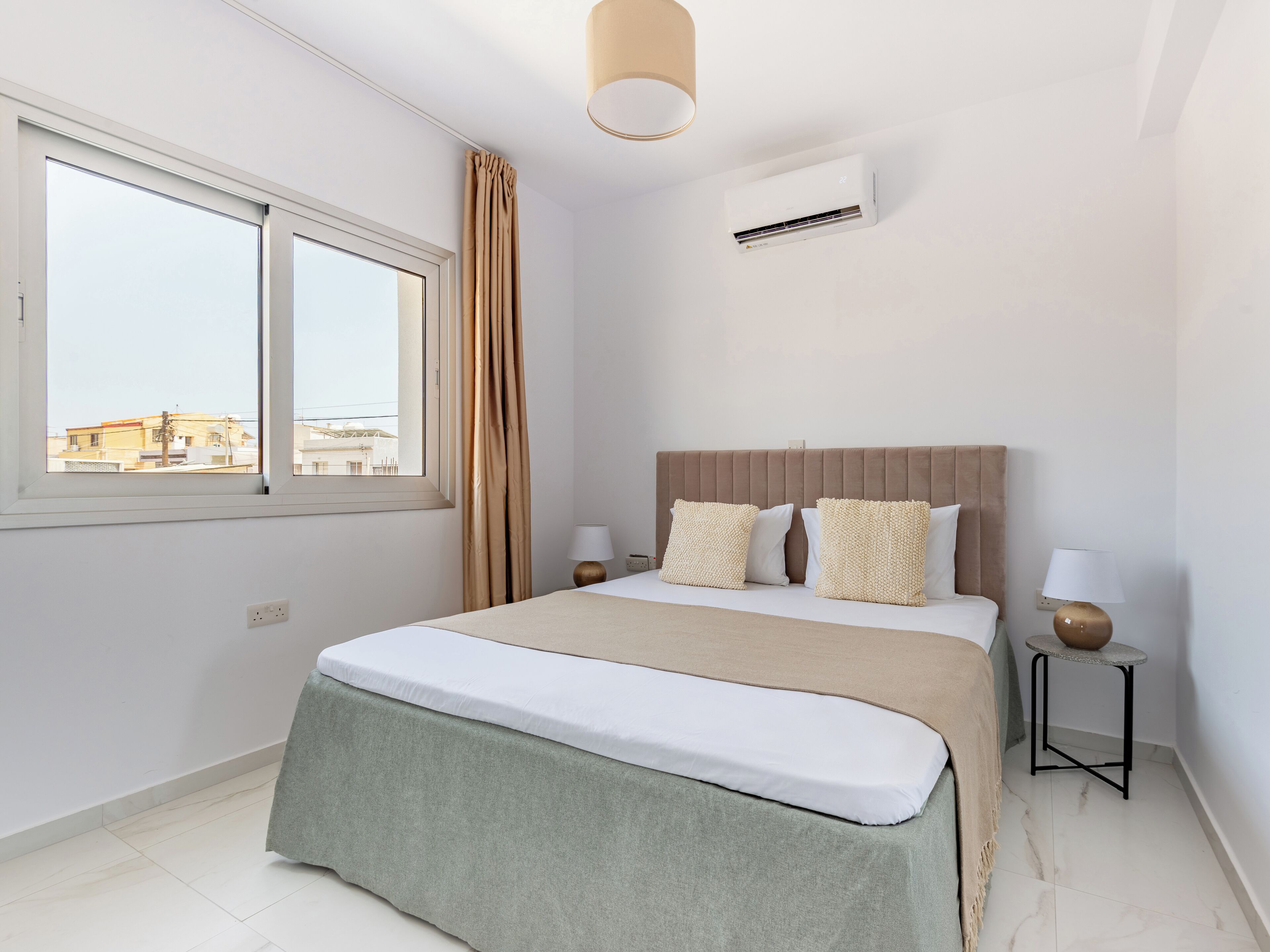 A hotel room with a bed, two bedside tables, a window, a curtain, an air conditioning unit, and a lamp.