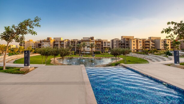 A modern residential complex with a central water feature, landscaped gardens, and a swimming pool. - Prime Select New Giza (6th of October City)