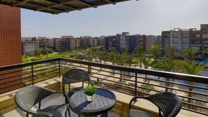 A balcony with wicker furniture and a potted plant, overlooking a residential area with palm trees and a swimming pool. - Prime Select New Giza (6th of October City)