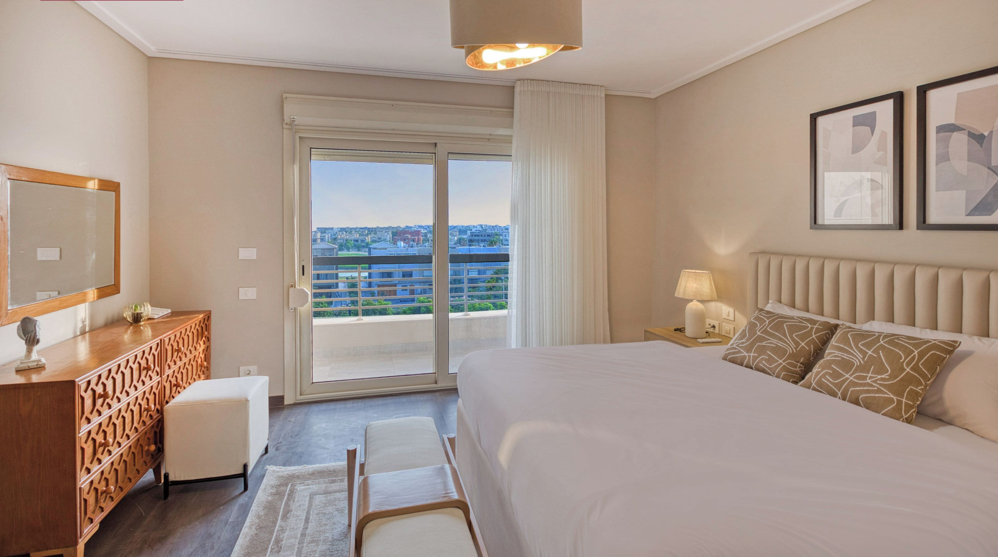 A hotel room with a large bed, a dresser, a mirror, a nightstand with a lamp, and a view of the city through the window.