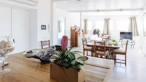 Zona d'estar àmplia amb taula de dinar de fusta, cadires i una televisió. Hi ha una planta sobre la taula i una perxa de roba al fons.