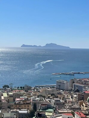 A coastal cityscape with buildings, a marina, and a clear view of the sea and distant mountains.