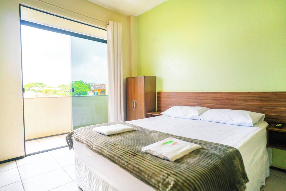 A hotel room with a large bed, a wardrobe, a nightstand, and a sliding glass door leading to a balcony.
