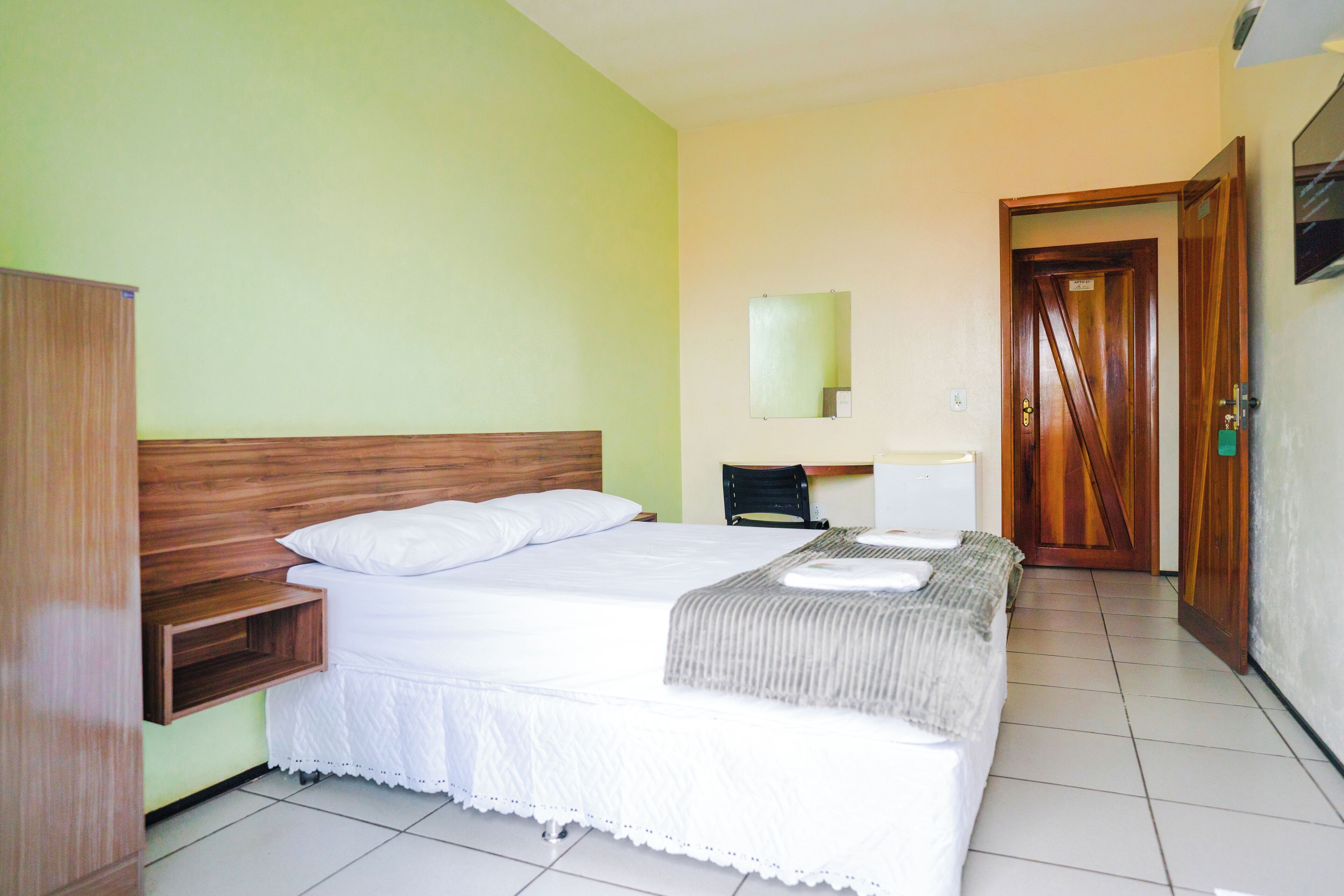 A hotel room with a wooden headboard, a bed with white bedding, a bedside table, a chair, a desk, and a mirror on the wall.