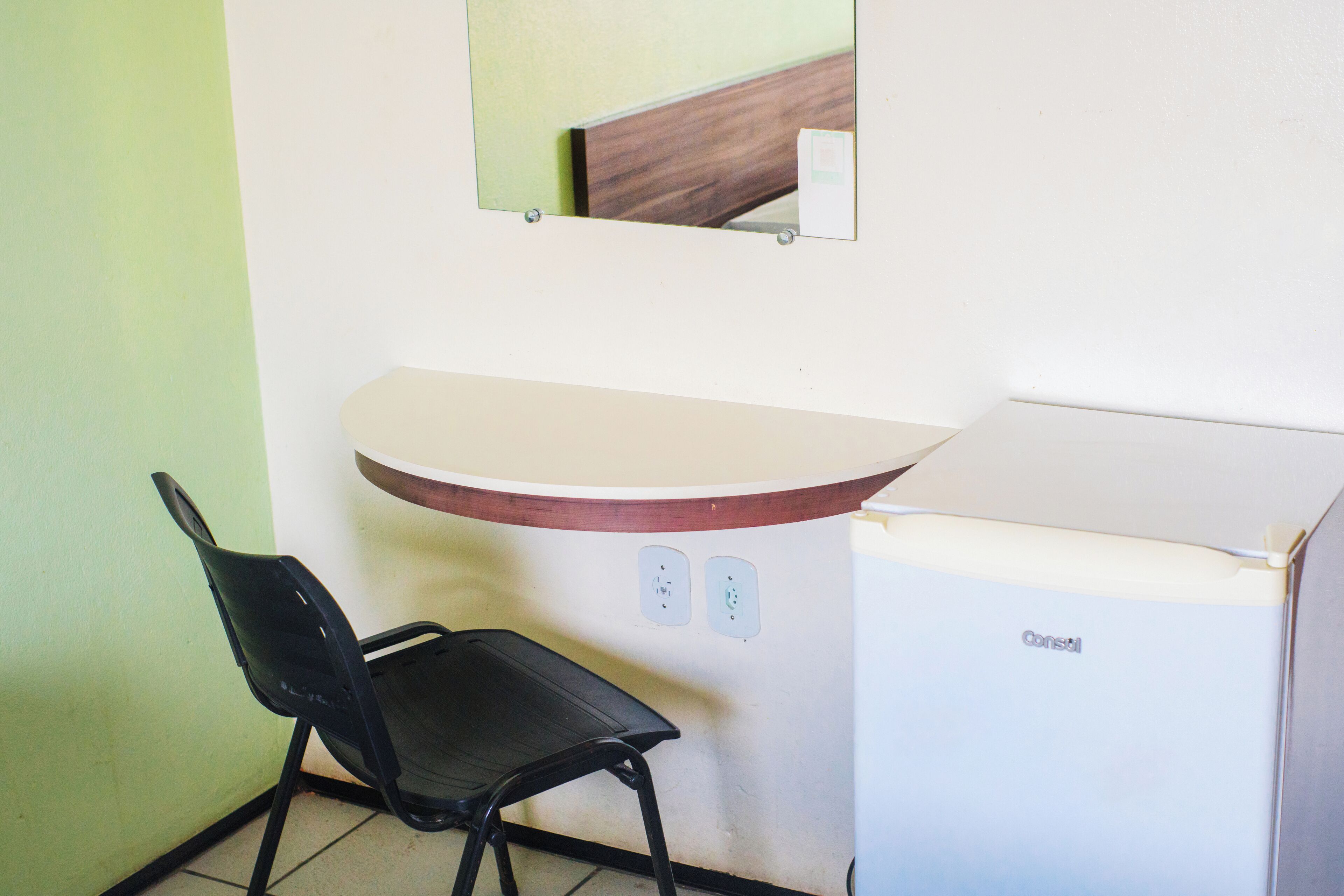 Une petite pièce avec un bureau blanc, une chaise noire, un miroir et un réfrigérateur.