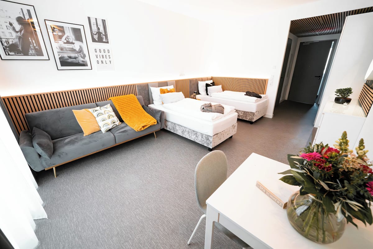 A modern hotel room with a grey sofa, two single beds, a small table, and a chair. There are framed pictures on the wall and a vase with flowers on the table.