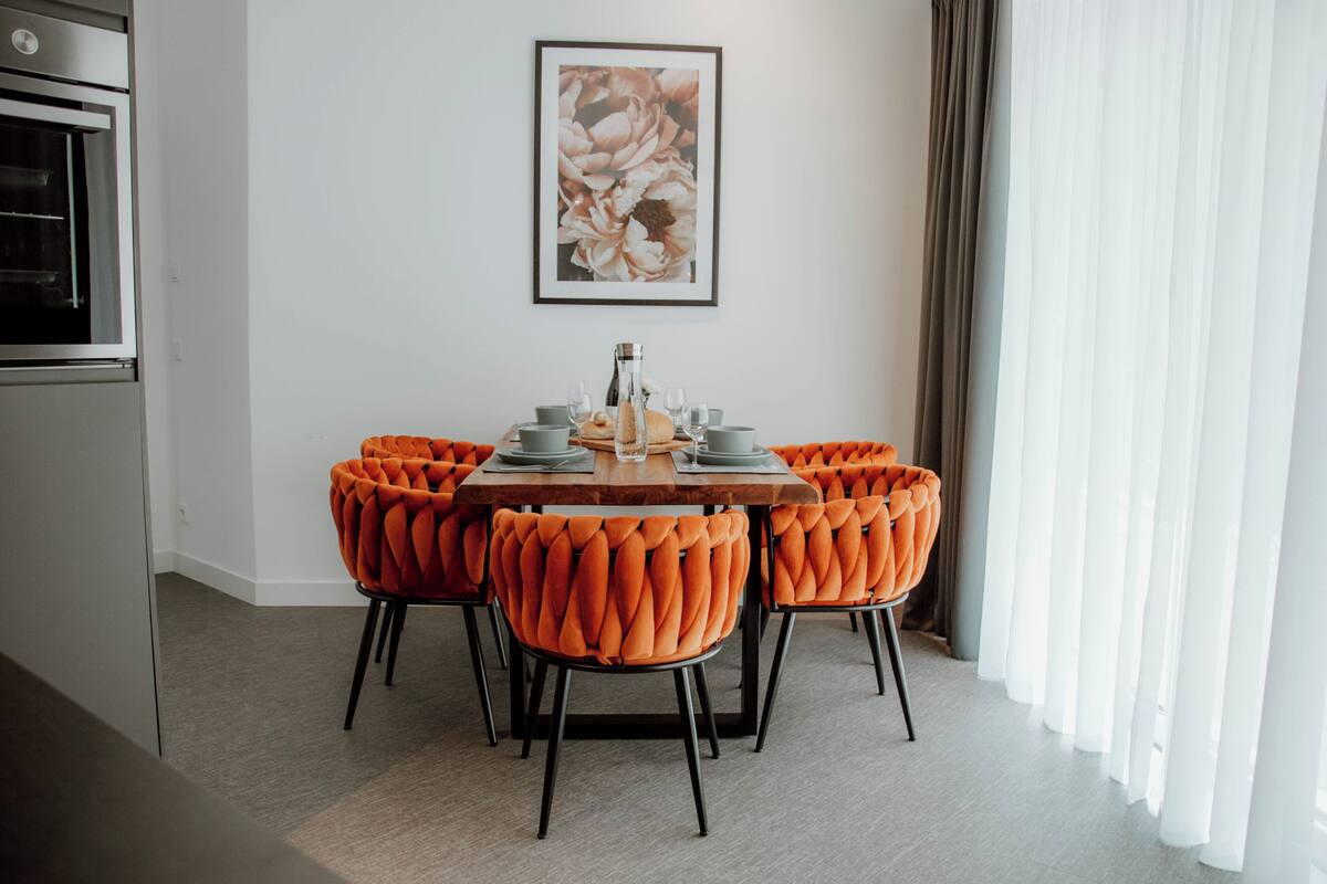 A dining area with a round table, orange upholstered chairs, and a framed floral artwork on the wall.