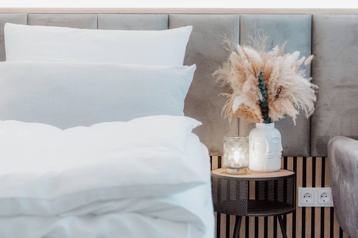 A neatly made bed with white linens, a bedside table with a vase of dried flowers, and a wall socket.