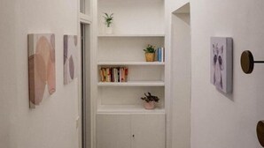 A narrow hallway with white walls and a wooden floor. There are shelves with books and plants, and a door on the left. - Pez's newly renovated house! (Roma)