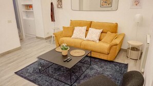A living room with a mustard-colored sofa, a coffee table with remote controls, and a blue rug. - Pez's newly renovated house! (Roma)