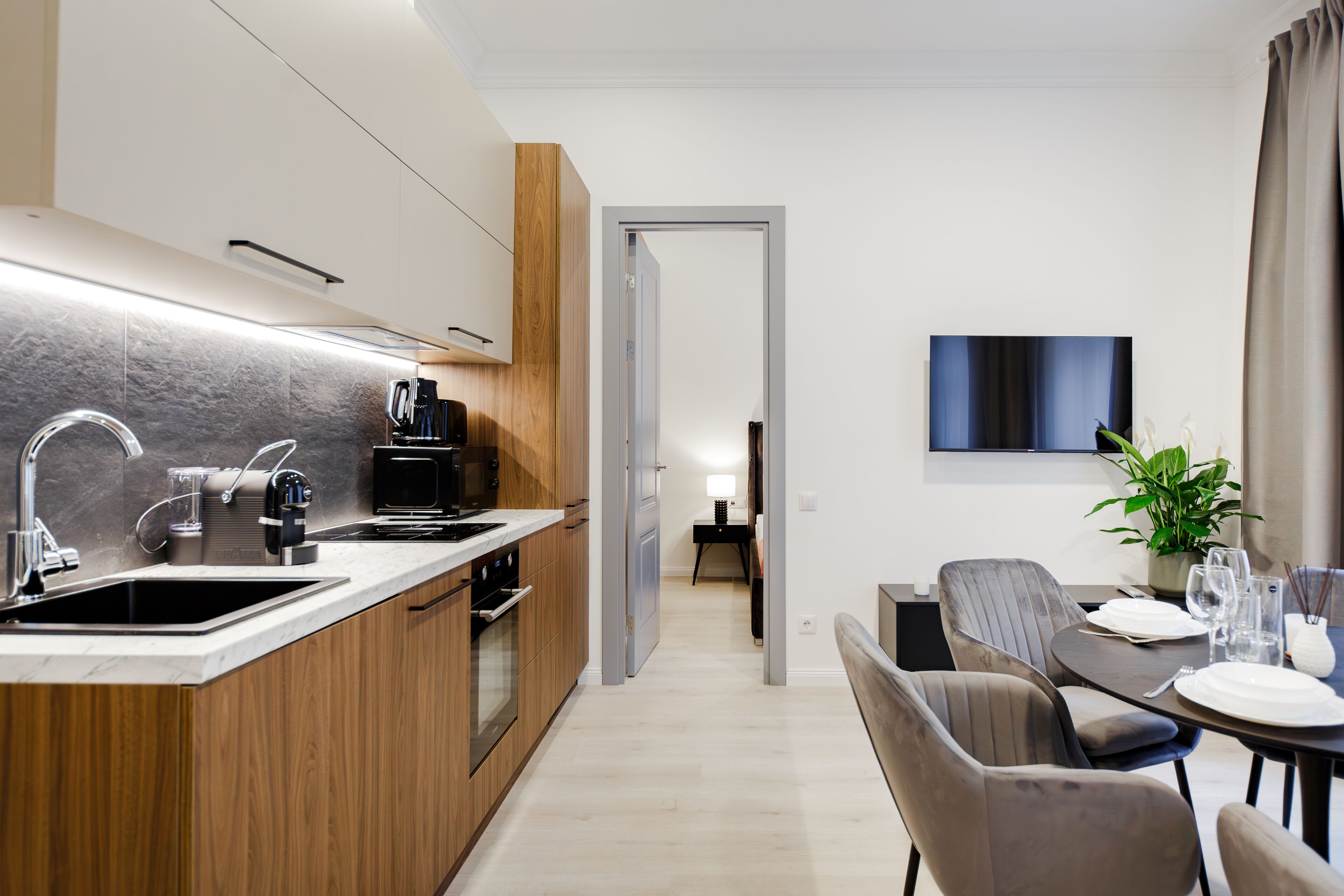 A modern kitchen with a built-in oven, a coffee machine, and a sink. There is a dining area with a table set for two and a plant on the table.
