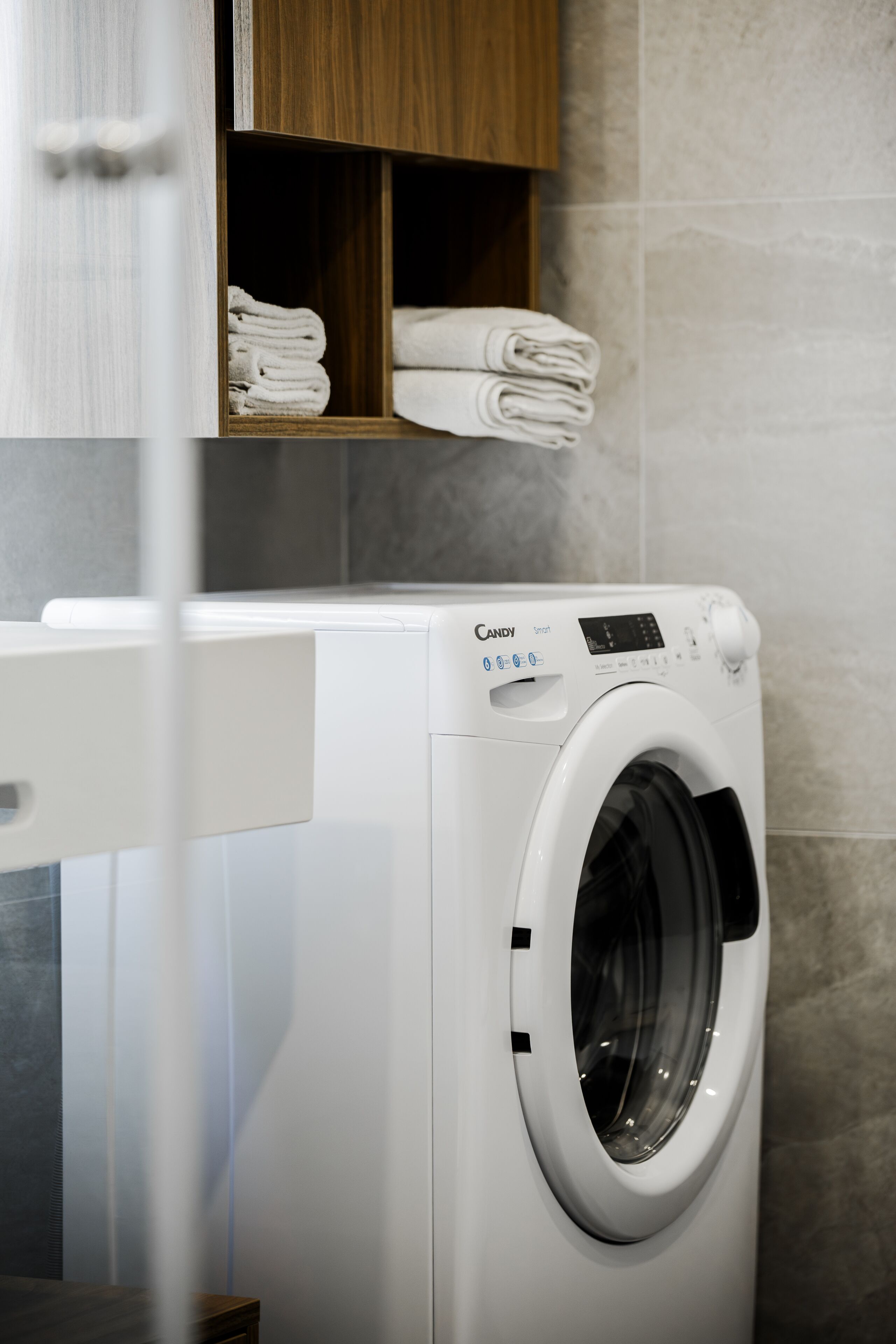 A modern laundry room with a white Candy Smart washing machine, wooden shelves with folded towels, and grey tiled walls.