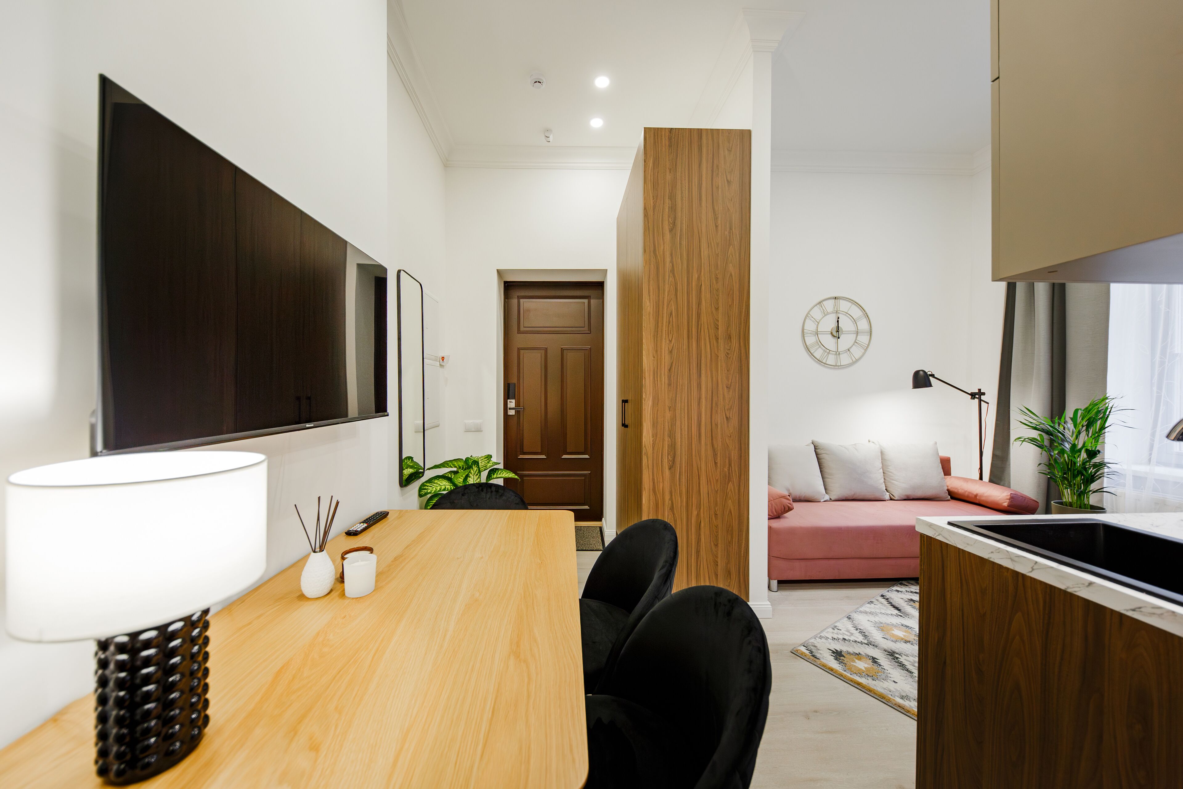 A modern living room with a wooden dining table, black chairs, a pink sofa, and a large flat-screen TV mounted on the wall.