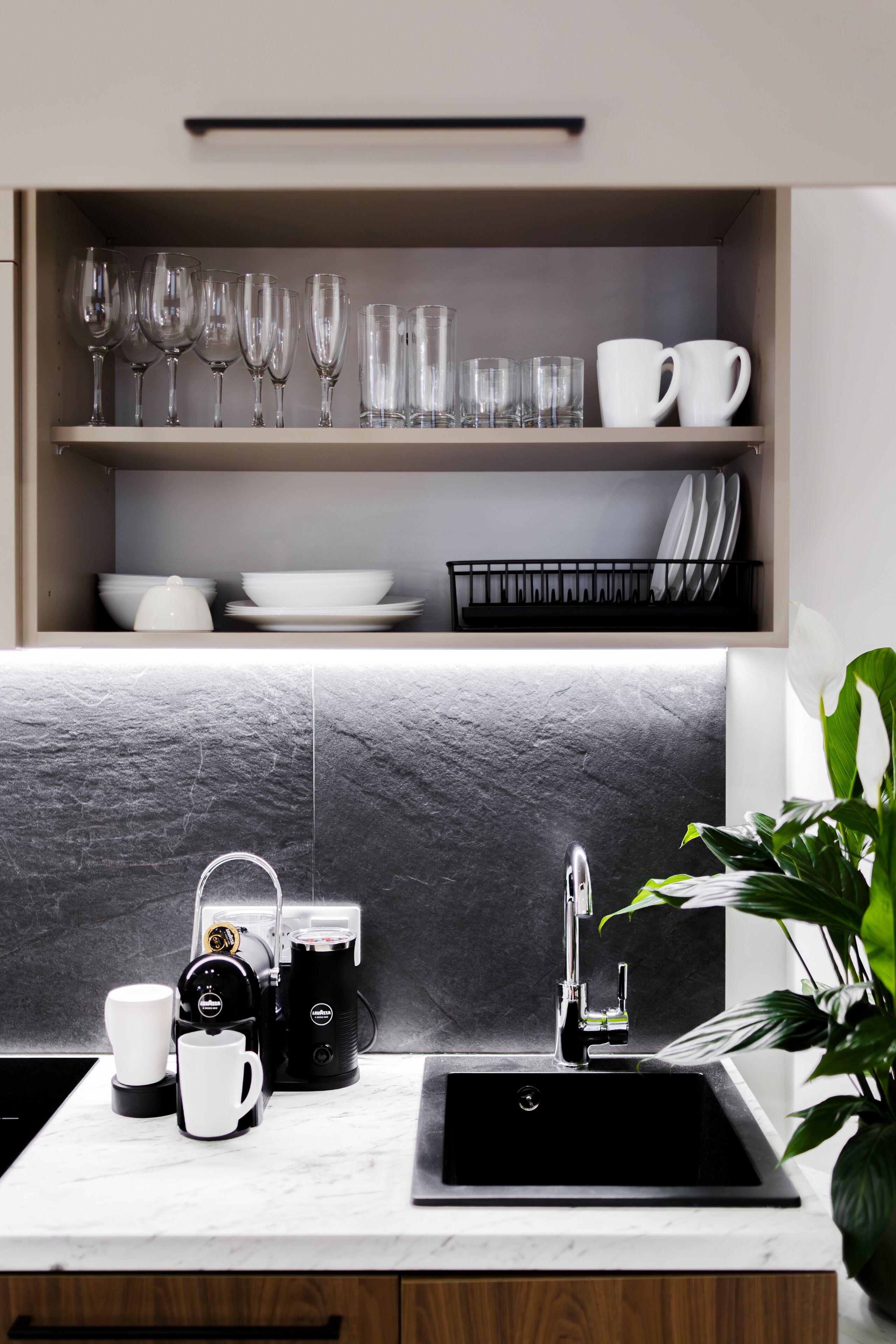 A modern kitchen with a marble countertop, a black sink, and a coffee machine. There are wine glasses and cups on the shelves above.