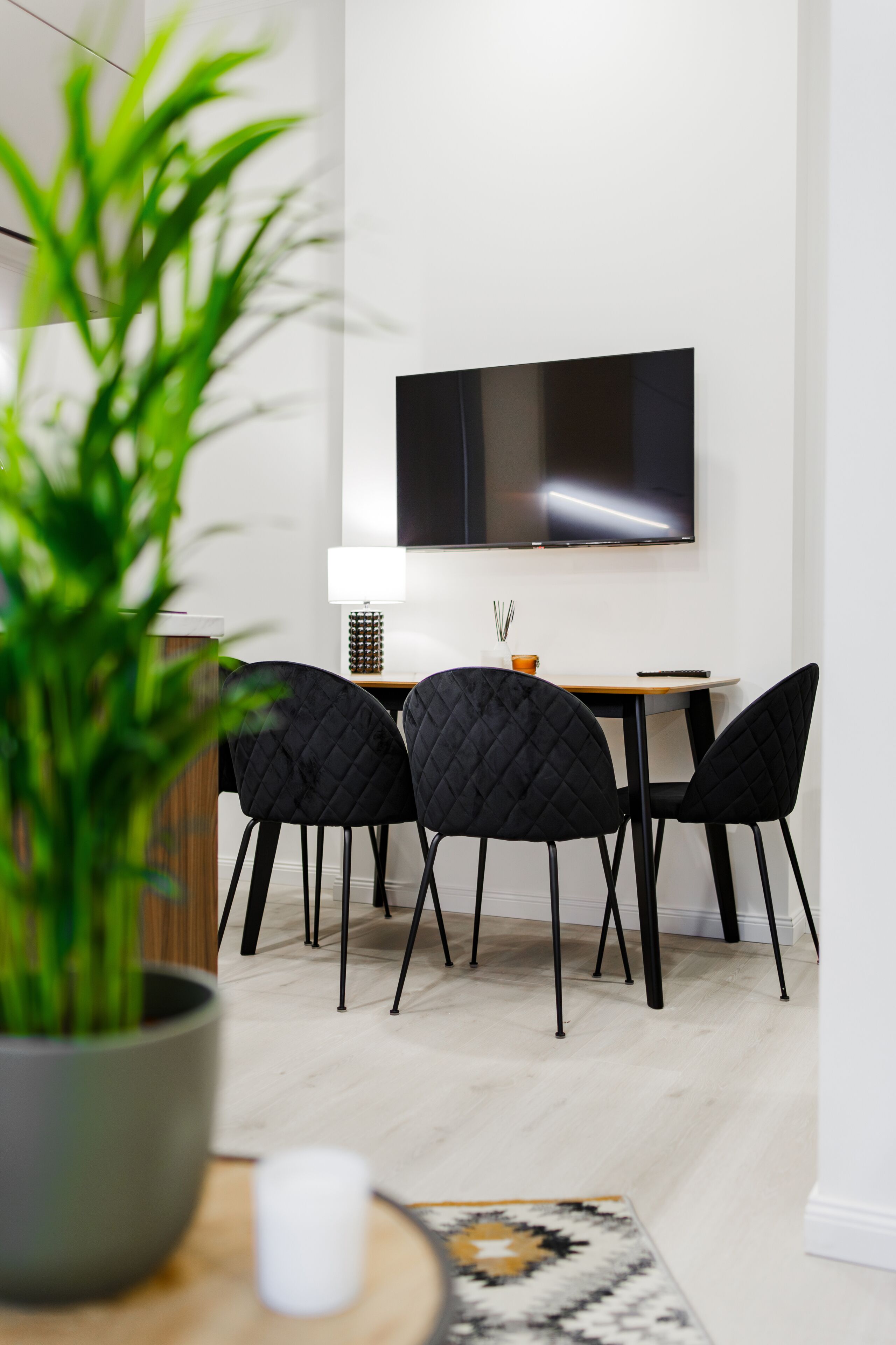 Un comedor moderno con un televisor montado en la pared, sillas negras, una mesa de madera y una planta en maceta.