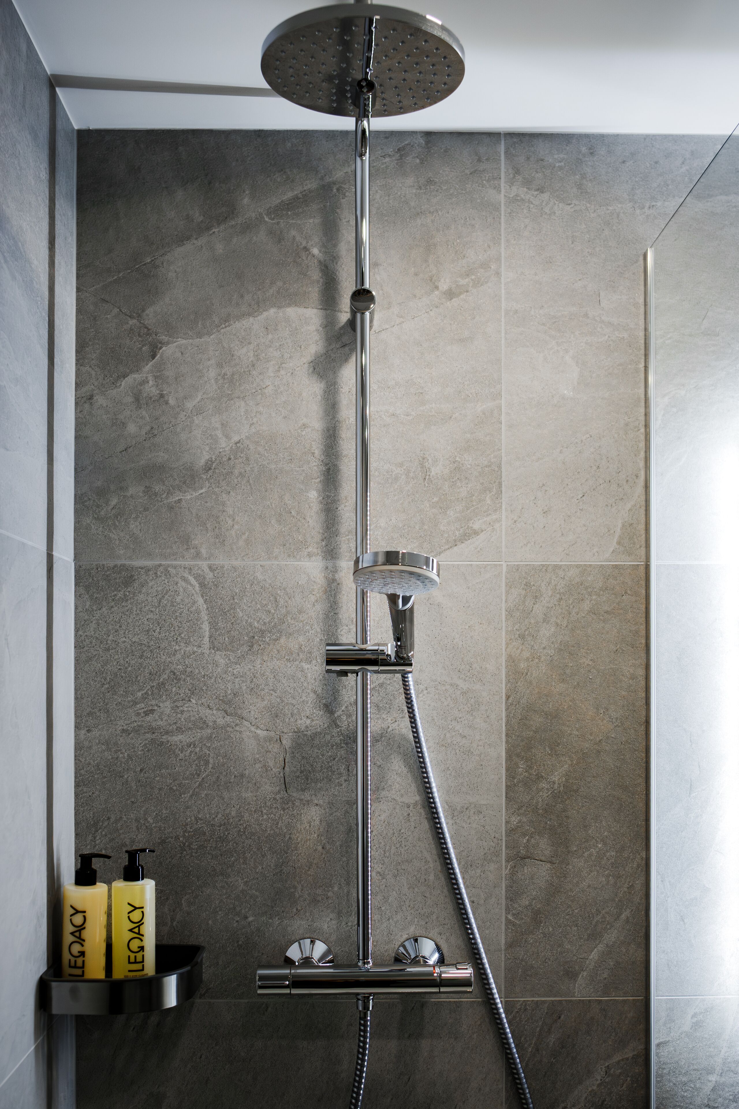 A modern shower with a rain showerhead and handheld showerhead, mounted on a wall with grey tiles.