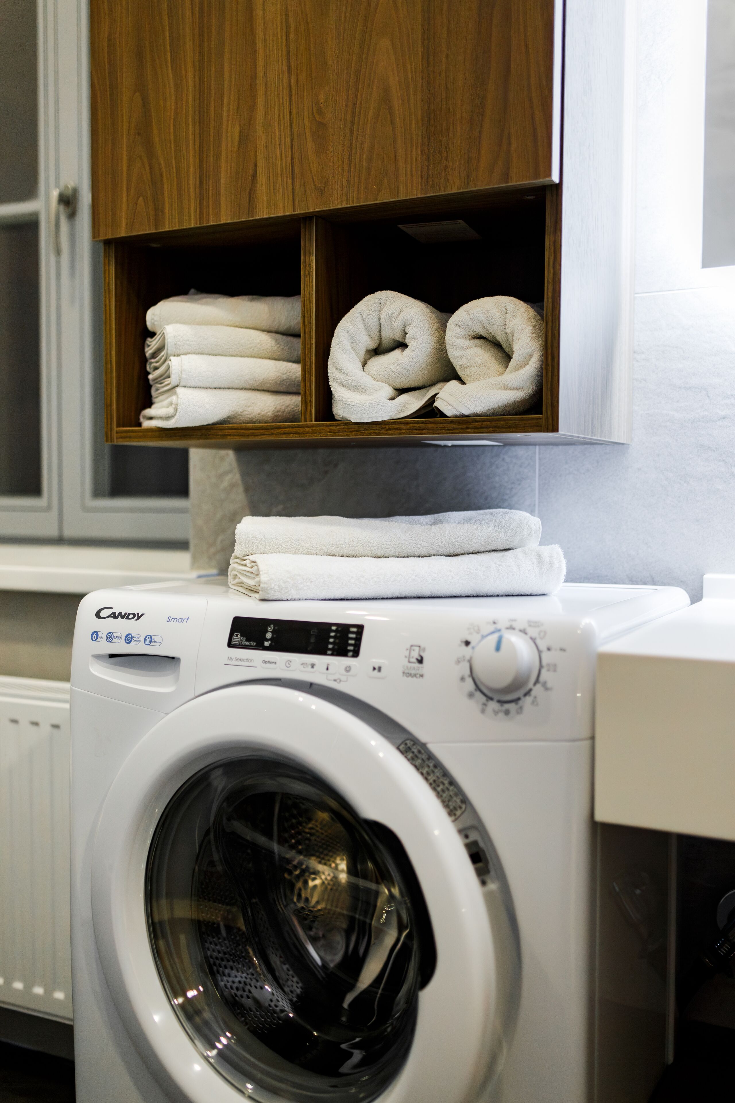 A front-loading washing machine with a control panel, a wooden shelf above it holding folded towels, and a white countertop to the right.