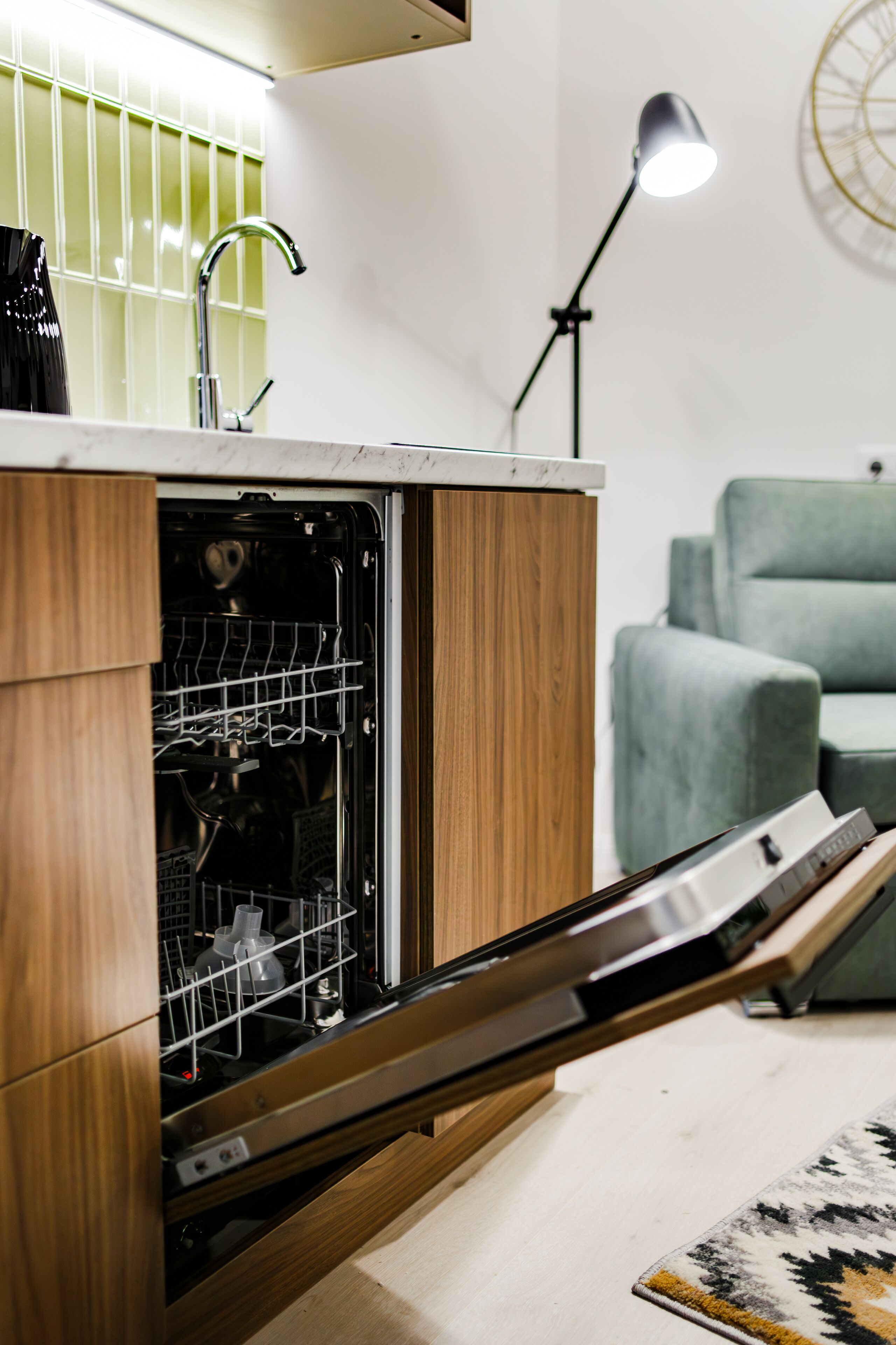A kitchen with an open dishwasher, a wooden cabinet, a green couch, and a floor lamp.