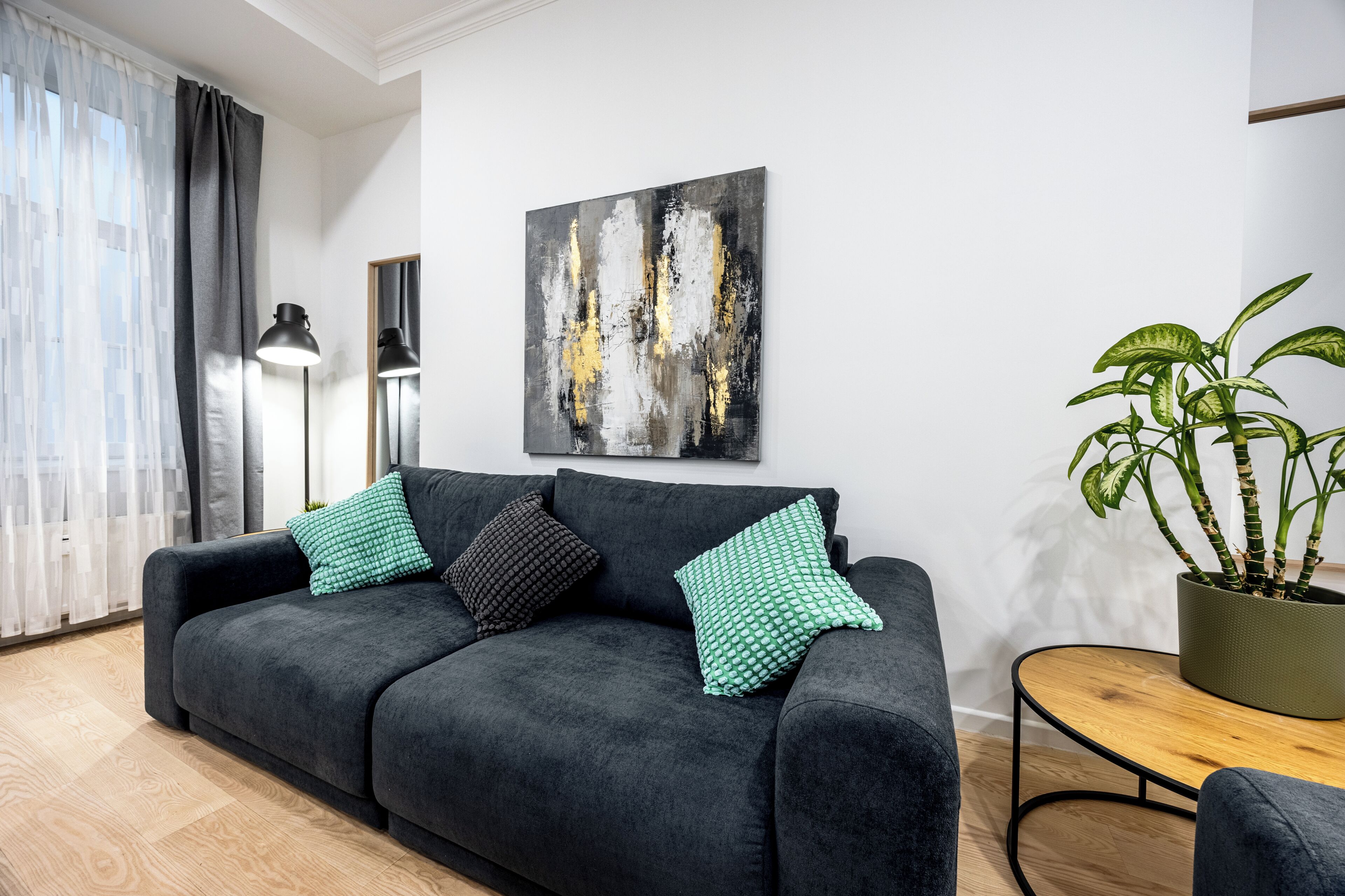 Ein modernes Wohnzimmer mit einem dunkelgrauen Sofa, einem kleinen runden Holztisch, einem Blumentopf und einem großen abstrakten Gemälde an der Wand.