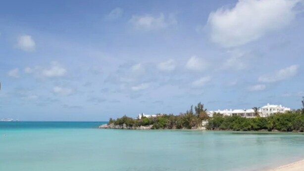 A coastal view with a beach, clear turquoise water, and a white building surrounded by greenery.