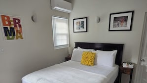 A neatly made bed with a white comforter and a yellow accent pillow, a small bedside table with a lamp, and a framed picture on the wall.