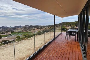 Een houten terras met een tafel en stoelen, uitkijkend op een glooiend landschap met velden en gebouwen in de verte.